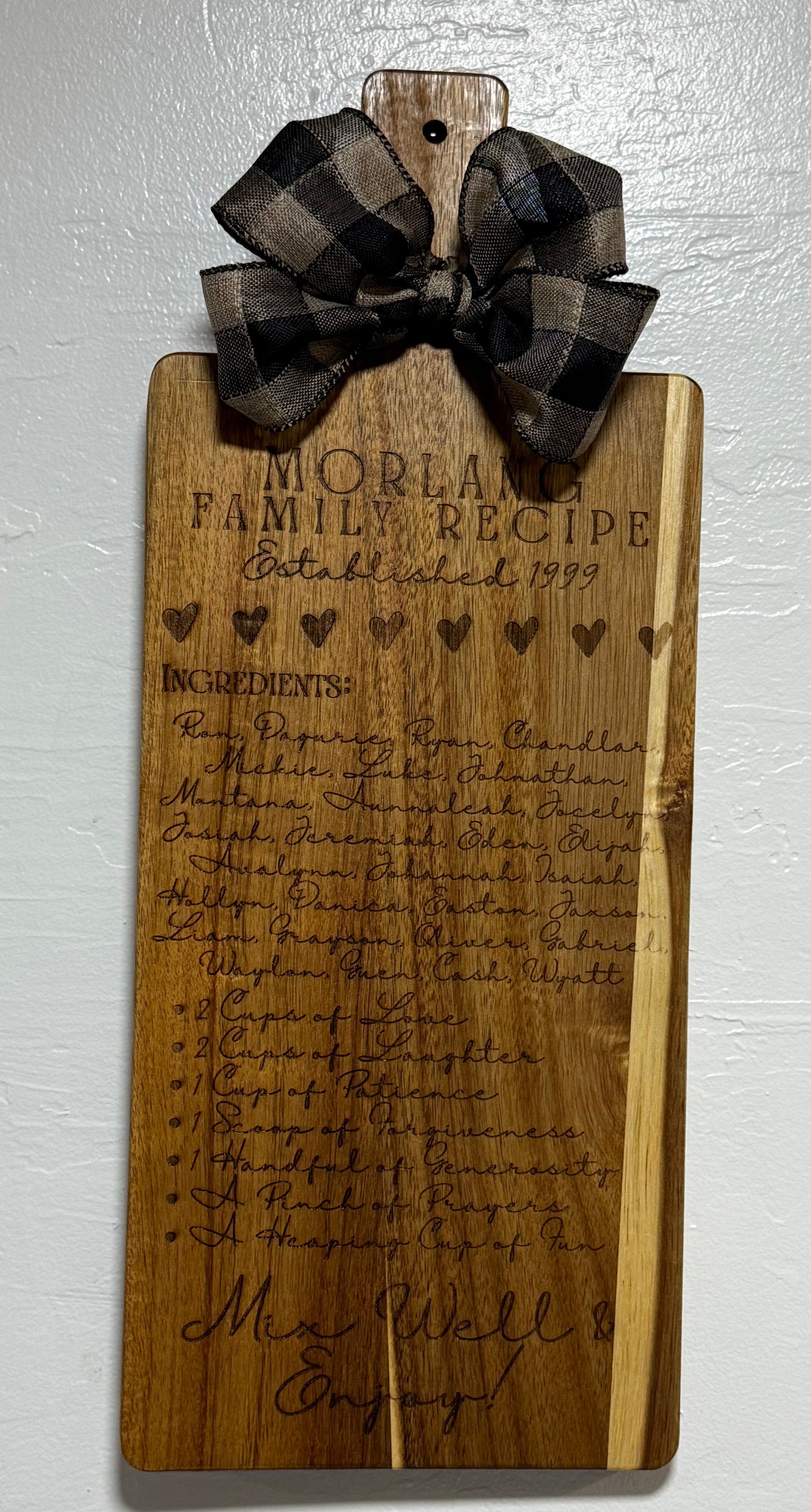 Wooden cutting board with engraved family recipe and a plaid ribbon on a white background